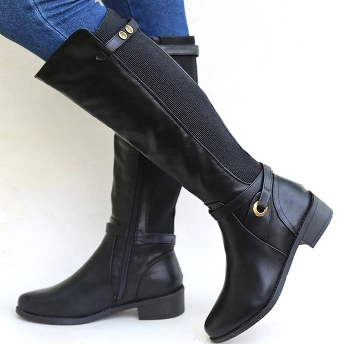 Botas altas negras con panel elástico frontal, detalles de hebilla y tacón bajo, combinadas con vaqueros ajustados.