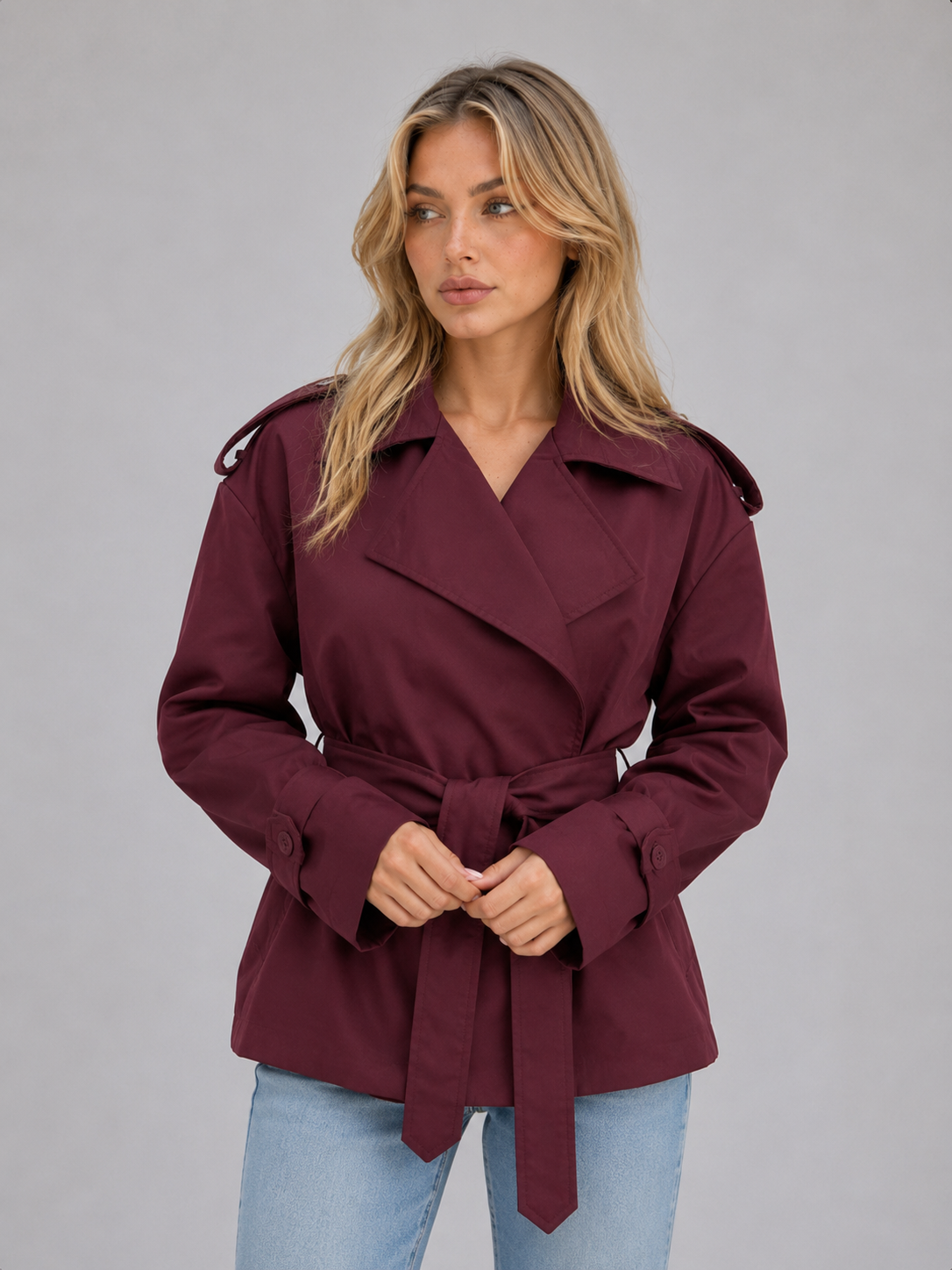 Gabardina corta color burdeos con solapas amplias, cinturón anudado al frente, hombreras abotonadas y puños con botones.