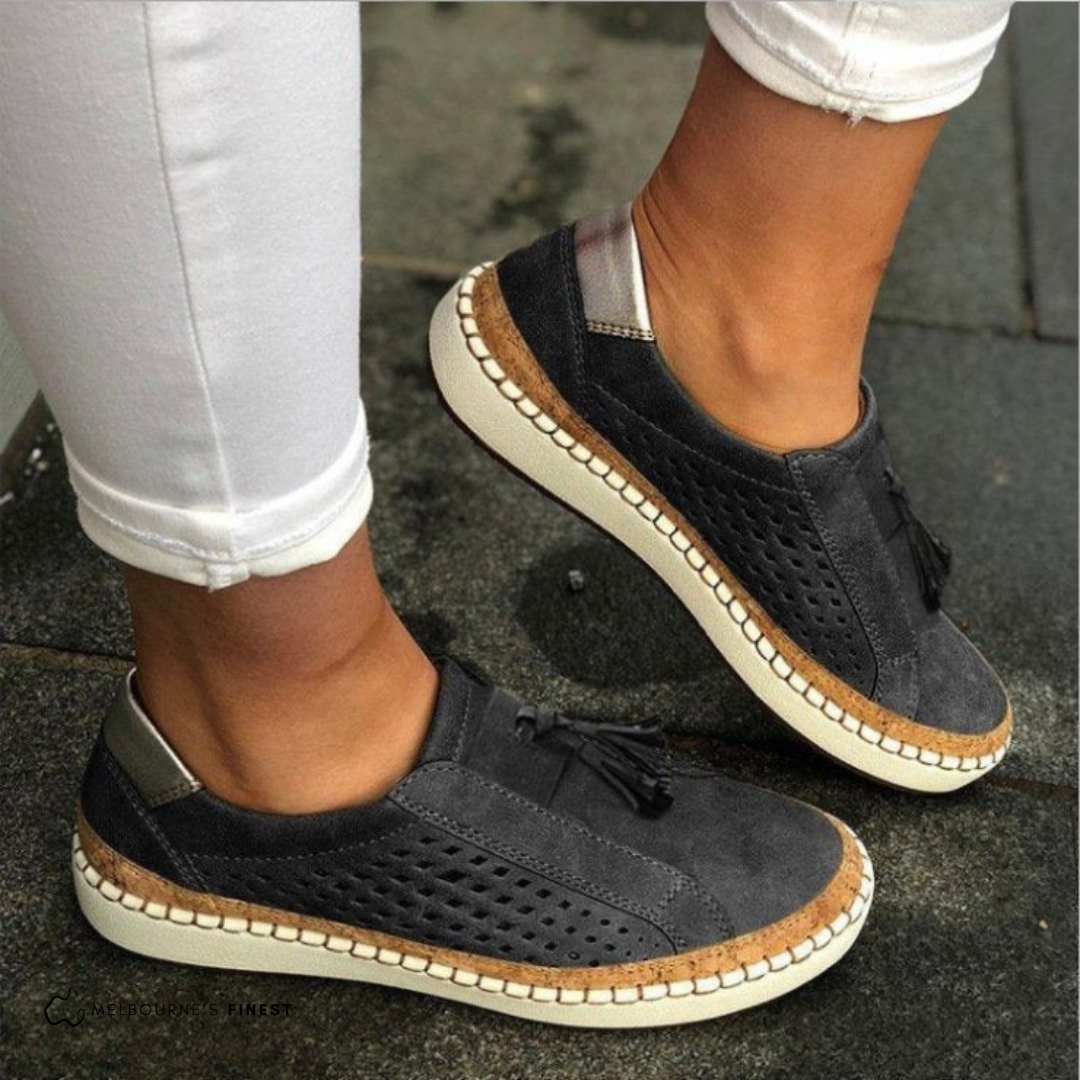 Mocasines planos negros con laterales perforados, borlas y suela clara, combinados con pantalón blanco.
