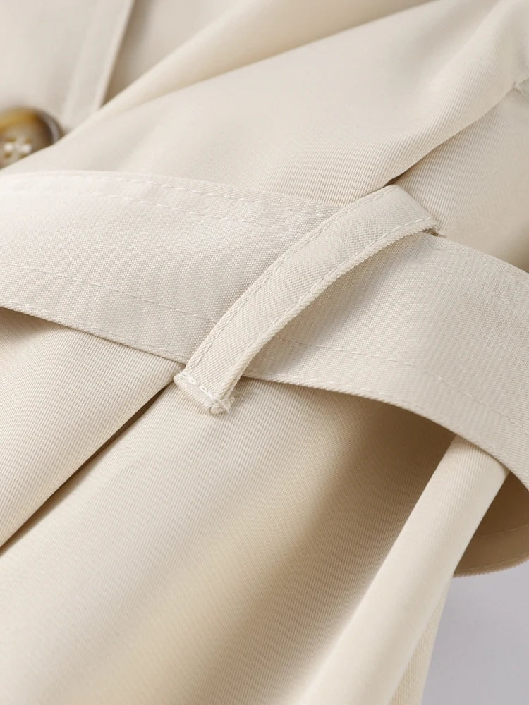 Acercamiento de una trabilla estrecha del trench beige sobre el tejido, con costuras y pliegues visibles.