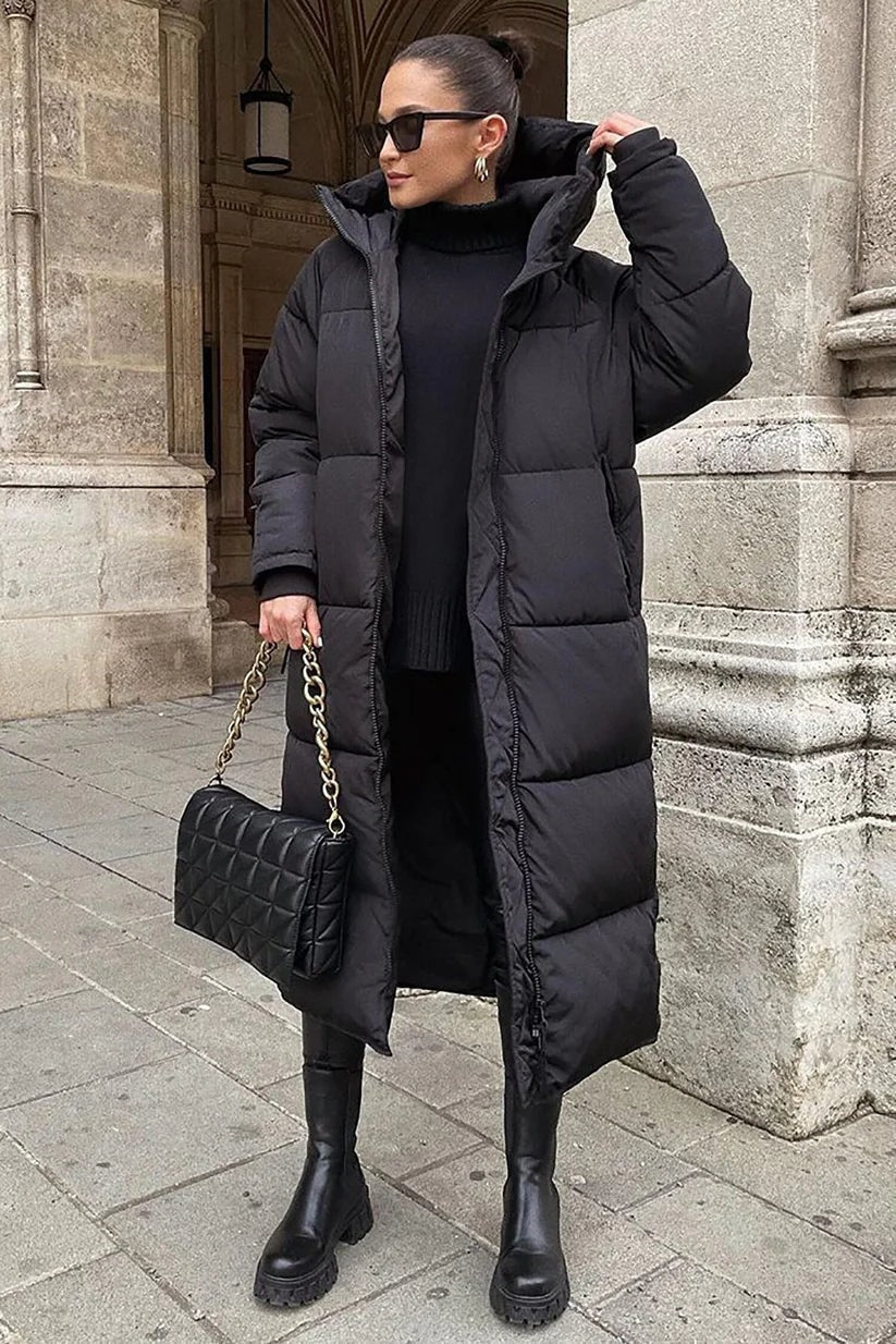 Abrigo acolchado largo negro con capucha, corte amplio y cierre frontal, combinado con ropa negra y botas altas.