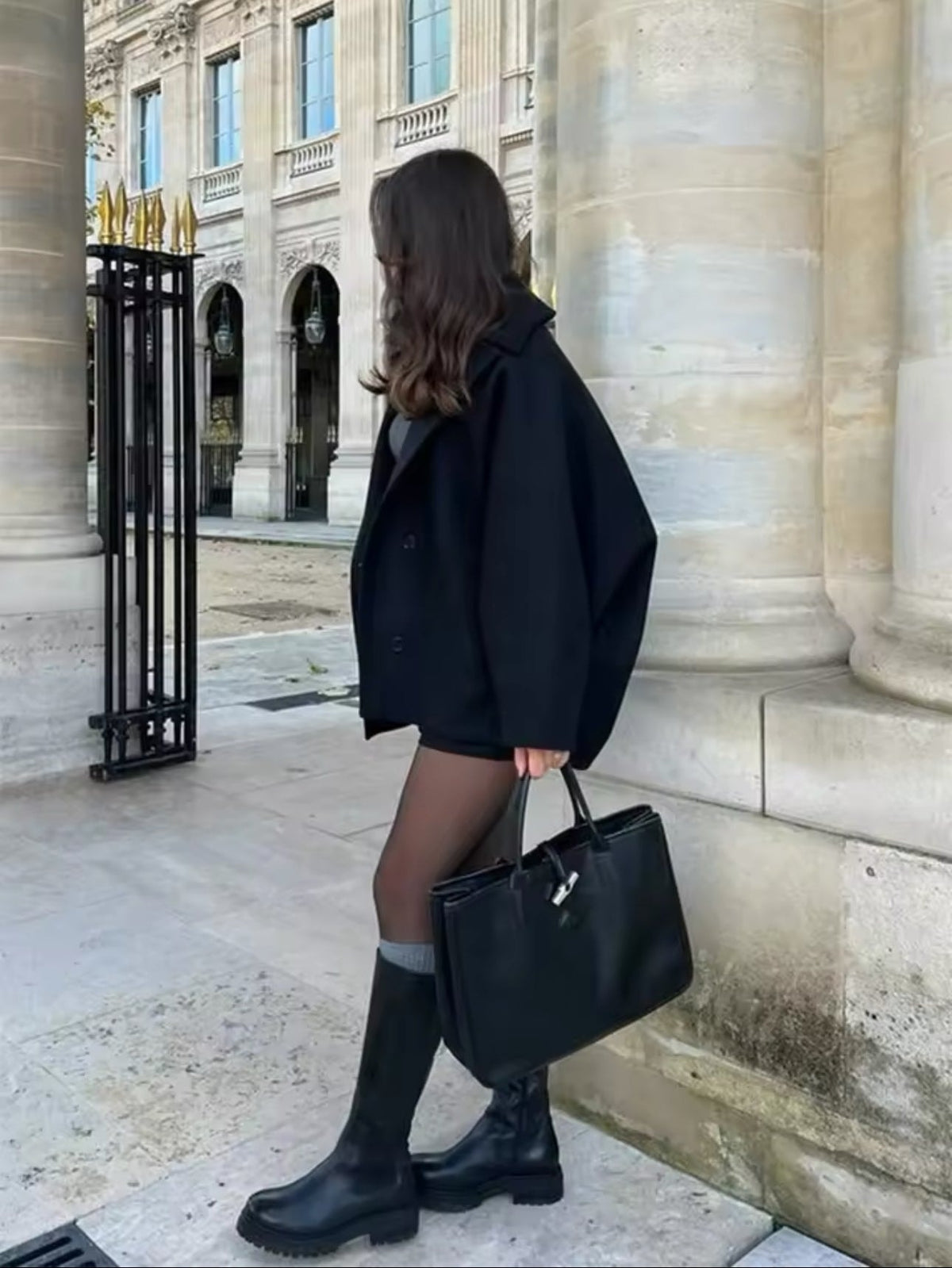 Vista lateral del abrigo negro abotonado, que muestra su silueta corta y recta mientras se lleva con botas altas.