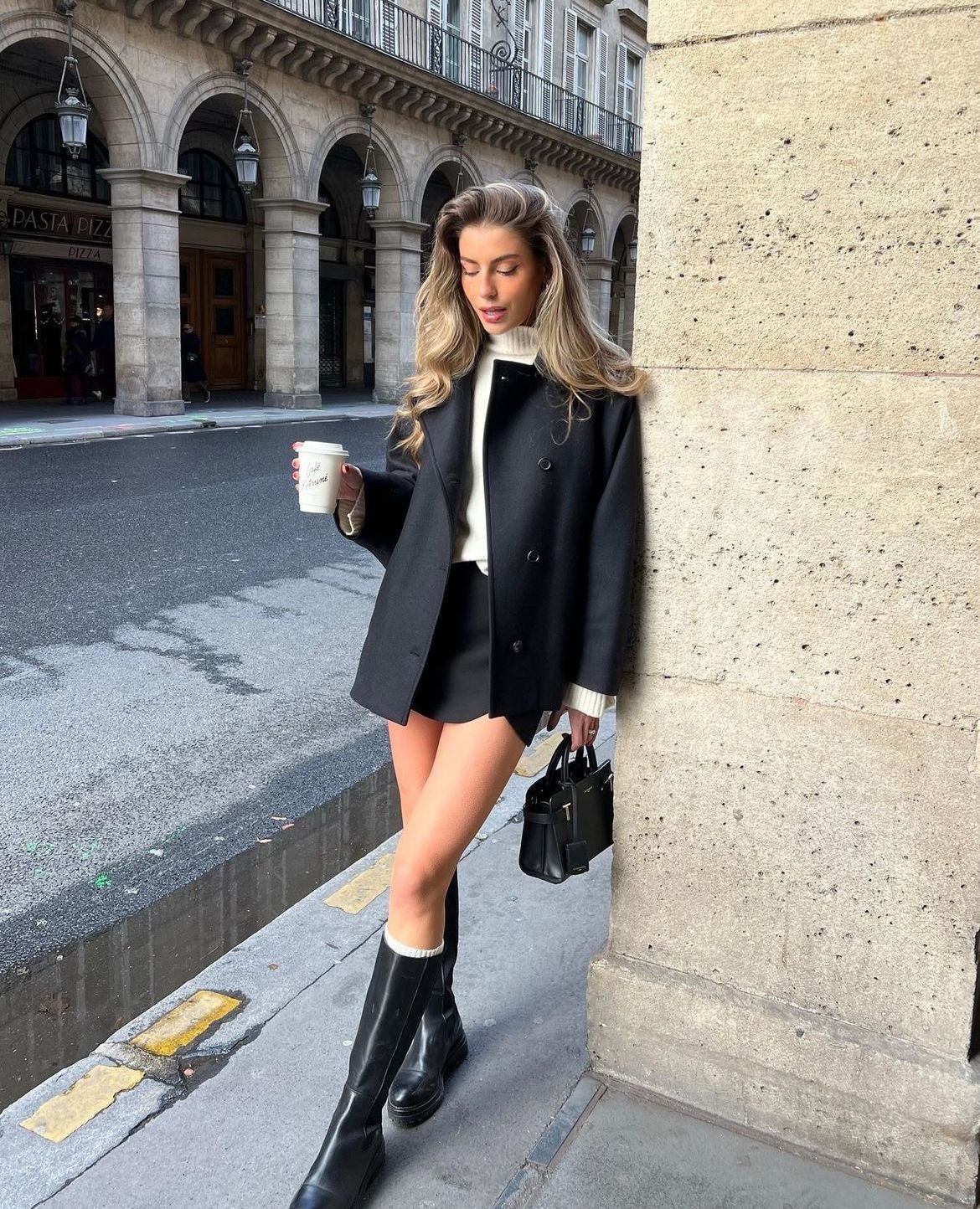 Abrigo negro corto de corte recto con cuello amplio y botones, combinado con prendas claras y botas en una calle soleada.