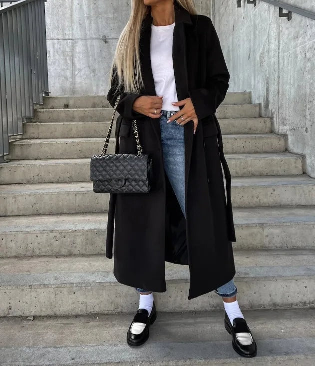 Abrigo largo negro abierto con solapas clásicas, combinado con vaqueros, camiseta blanca y mocasines.