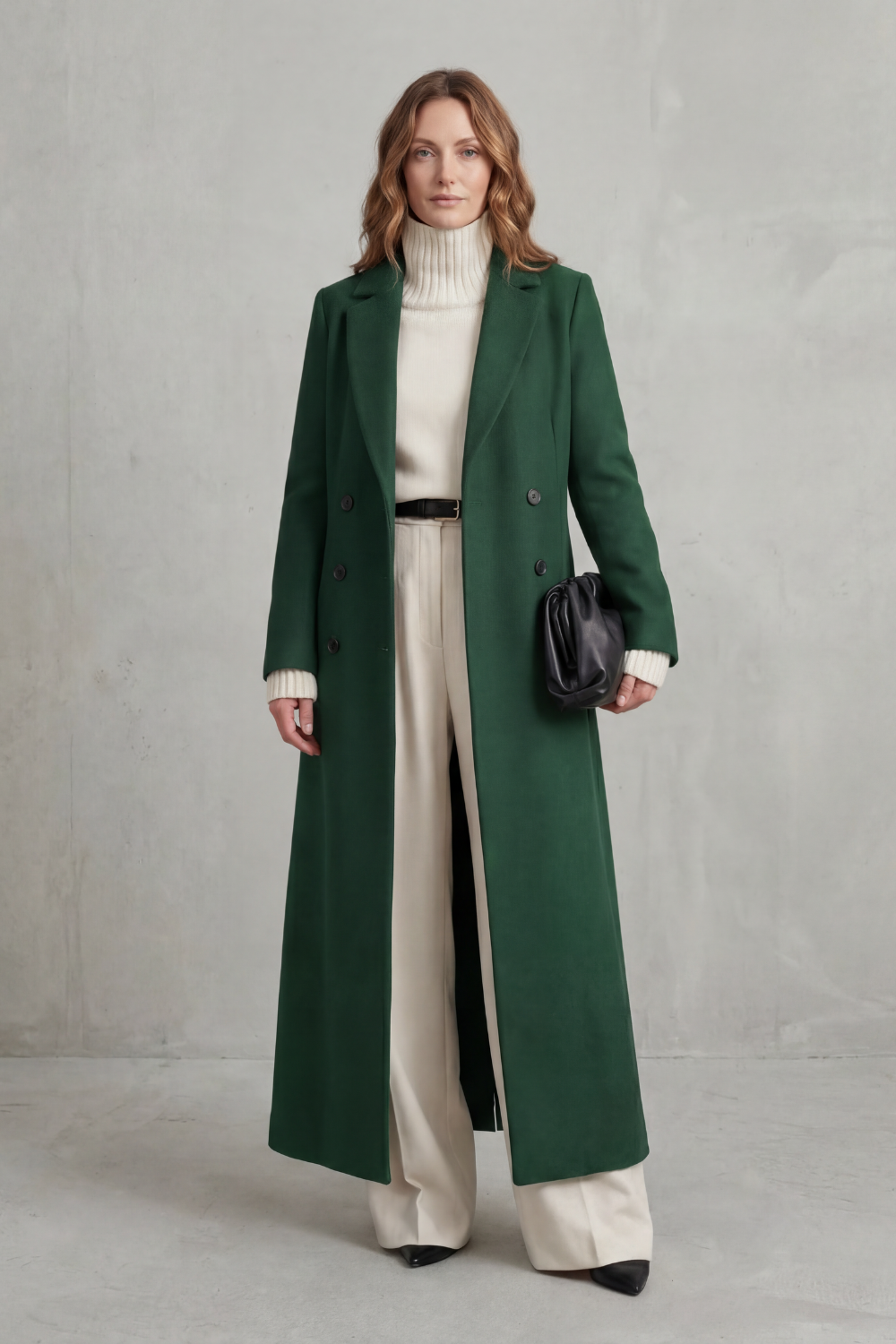 Vista frontal del abrigo largo verde oscuro, combinado con cuello alto claro, pantalones amplios y bolso negro.