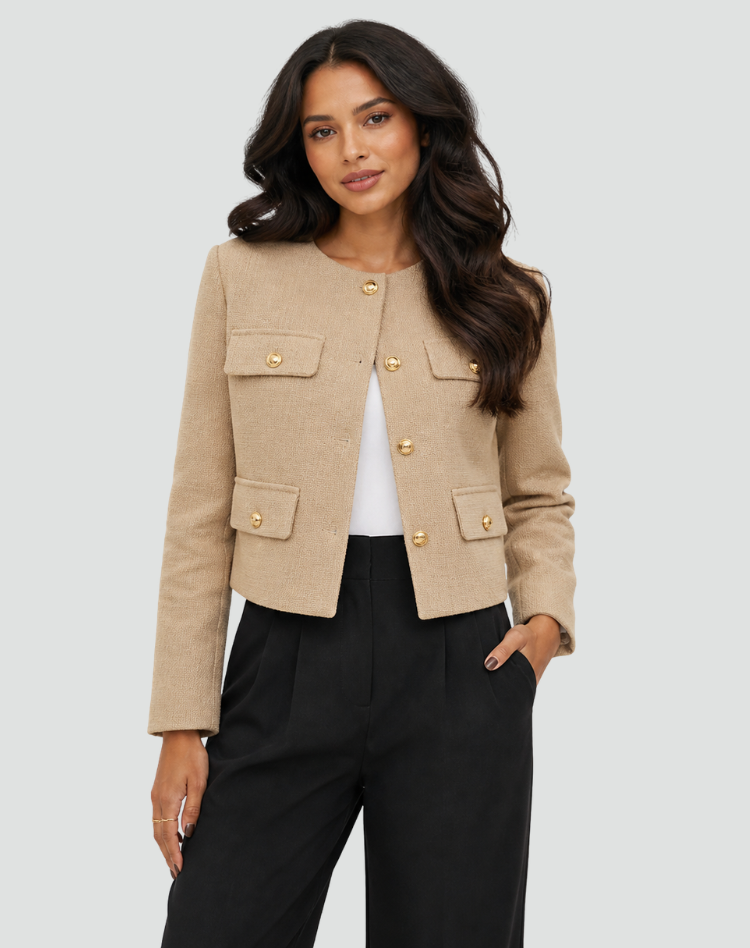 Blazer corto beige claro abierto sobre una prenda blanca, con botones dorados y bolsillos con solapa.