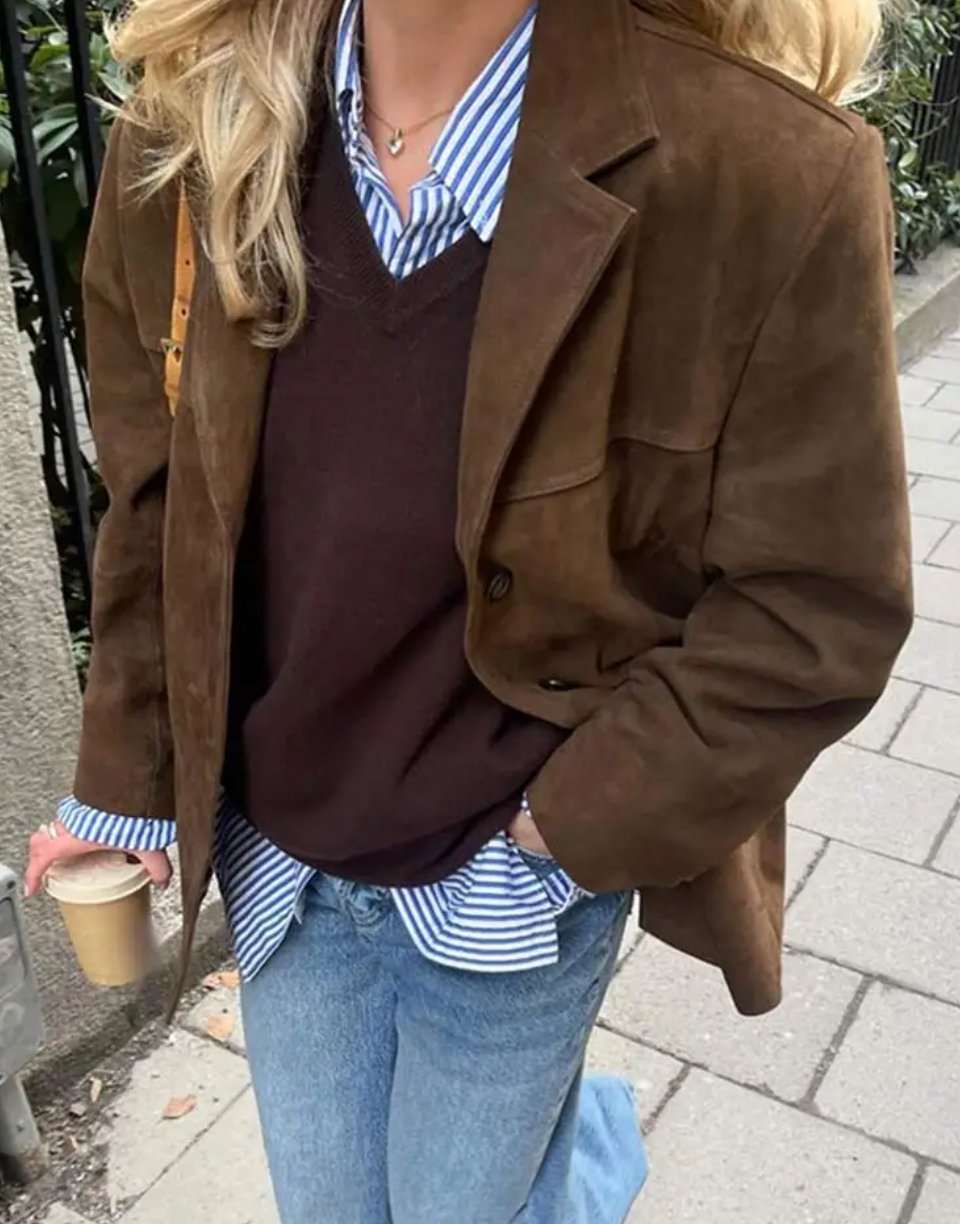 Blazer marrón de corte relajado con bolsillo en el pecho y solapas marcadas, combinado con camisa de rayas y vaqueros.
