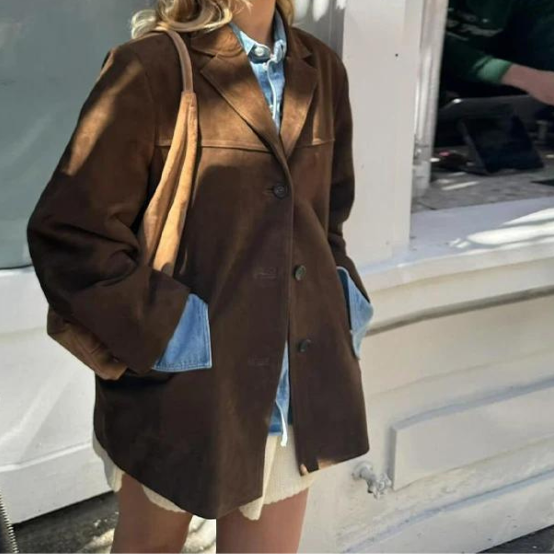 Blazer marrón de largo medio con cierre frontal de botones y bolsillos laterales, combinado con camisa vaquera y falda clara.