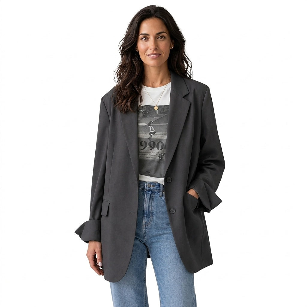 Blazer oversize gris oscuro con solapas clásicas, dos botones frontales y bolsillos, combinado con camiseta estampada y vaqueros.