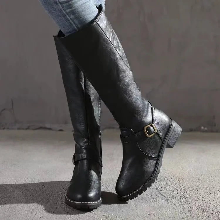 Botas altas negras con hebilla lateral, tacón bajo y suela dentada, mostradas en una vista cruzada con vaqueros.