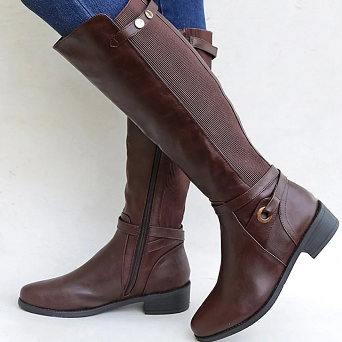 Botas altas marrones con panel elástico, hebilla decorativa y tacón bajo, mostradas sobre un fondo claro.