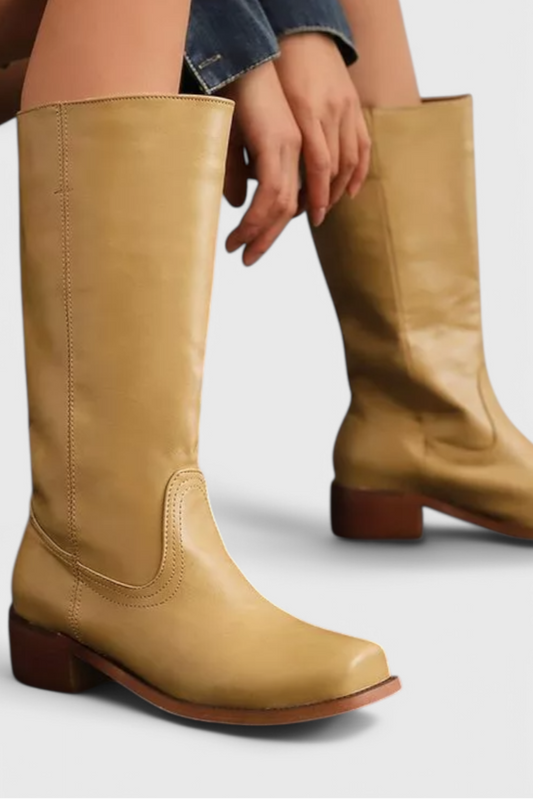 Bota alta color mostaza con tacón bajo, punta redondeada y costuras decorativas, junto a otra bota del mismo estilo.