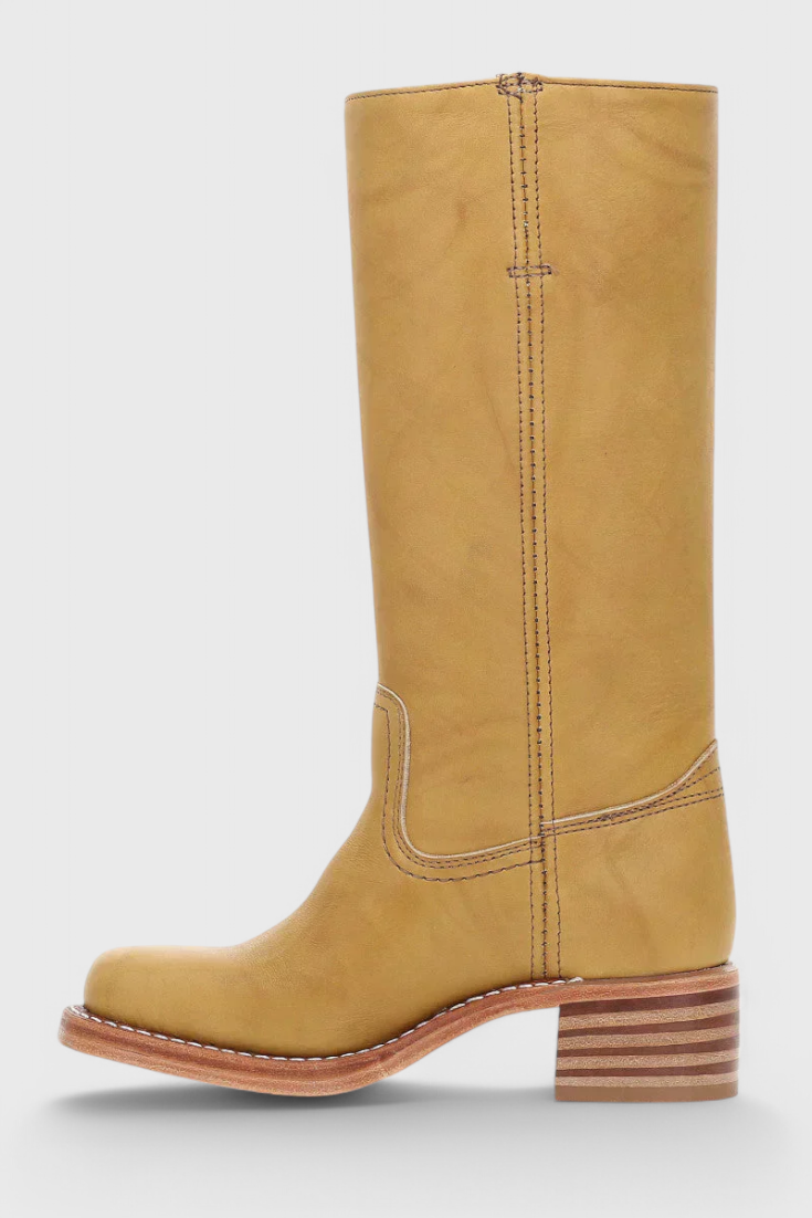 Bota alta mostaza en perfil lateral, con caña lisa, puntera redondeada y tacón bajo de capas horizontales.