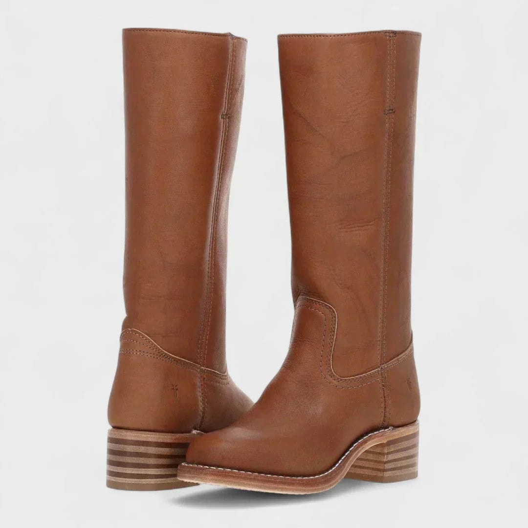 Botas altas color coñac de silueta ecuestre, mostradas cruzadas para destacar la puntera, la caña y el tacón bajo a rayas.