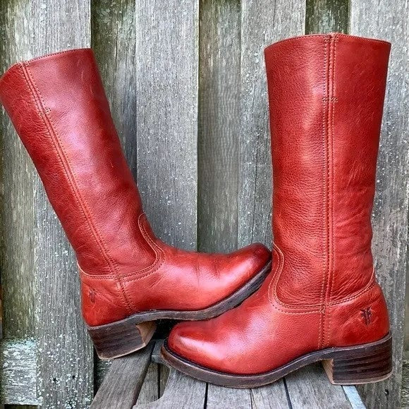 Botas altas rojas de estilo ecuestre sobre un fondo de madera, con una bota apoyada de lado y la otra erguida.