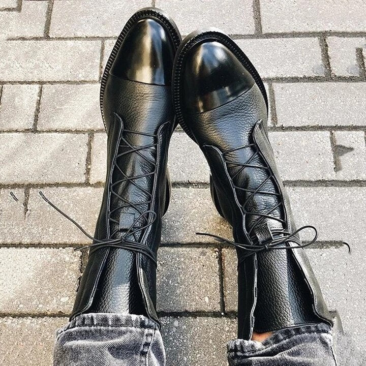 Botines negros con cordones cruzados y punteras brillantes, colocados sobre adoquines junto a unos pantalones vaqueros.