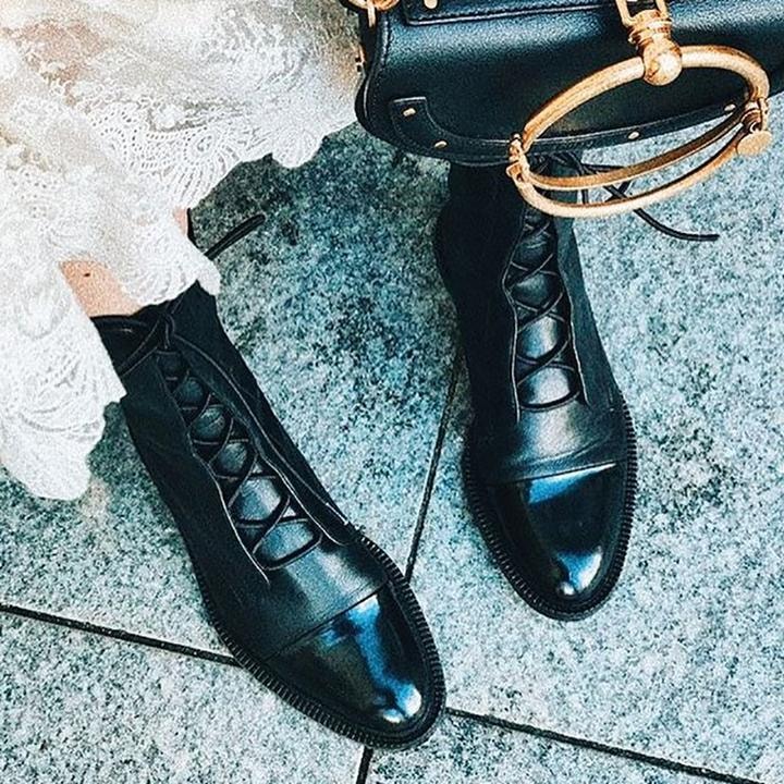 Botines negros con punteras brillantes combinados con una prenda blanca y un bolso negro sobre una superficie gris.