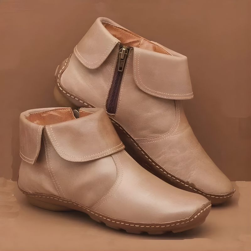 Botines planos beige dispuestos en ángulo, con solapas envolventes, costuras decorativas y cremallera lateral claramente visible en el modelo posterior.