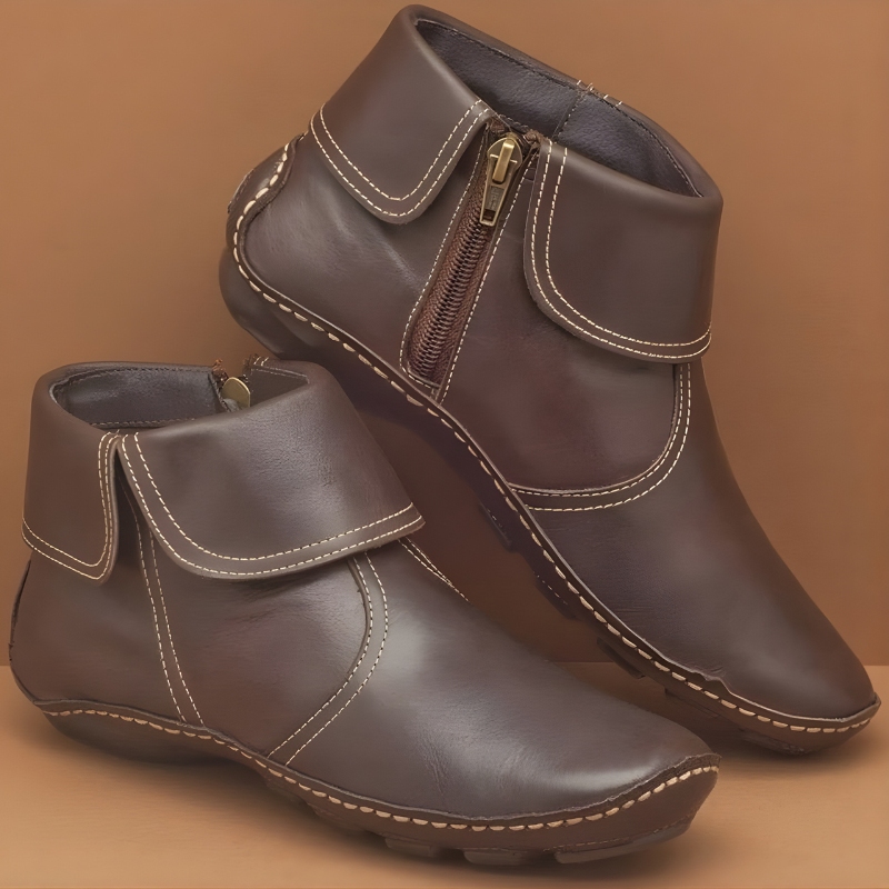 Botines planos marrón oscuro con suela dentada, solapas envolventes ribeteadas y costuras claras; uno aparece elevado detrás del otro.