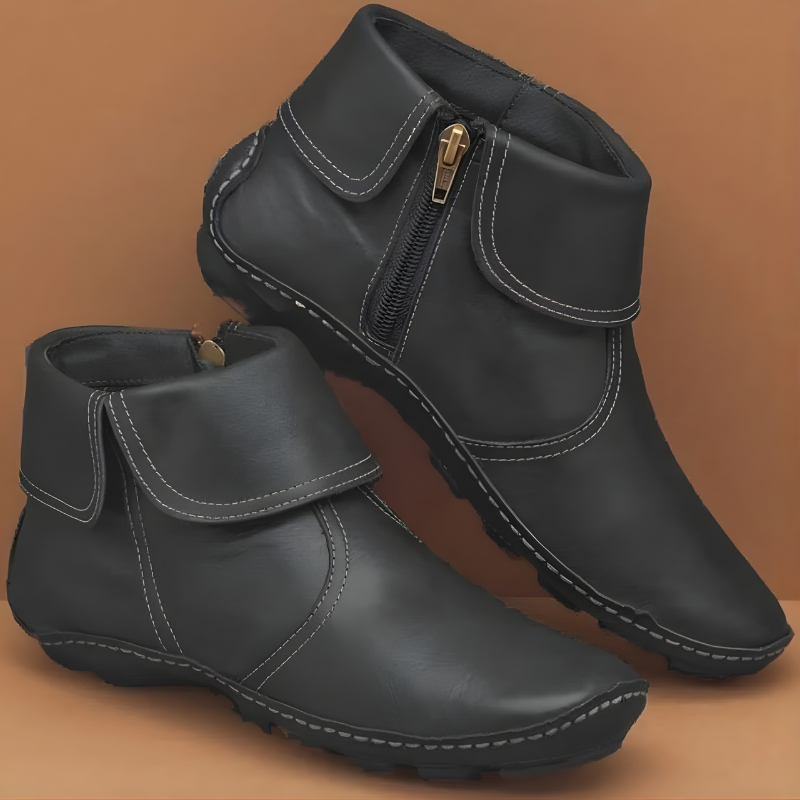Botines planos negros con contraste de costuras, cremallera lateral dorada y detalle envolvente; el par se presenta sobre un fondo marrón.