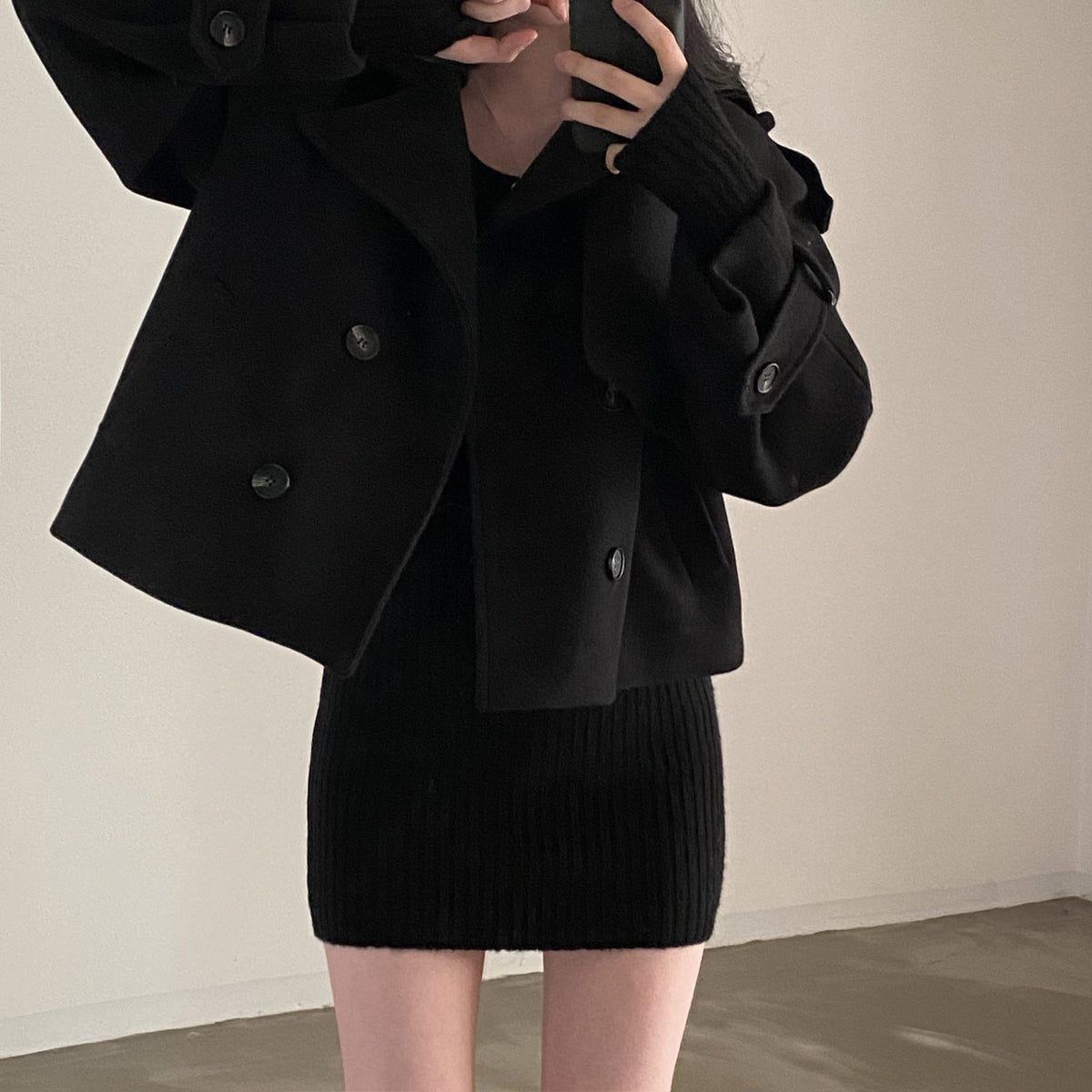 Caban corto negro de doble botonadura, combinado con un vestido de punto negro y mostrado en una vista frontal parcial.