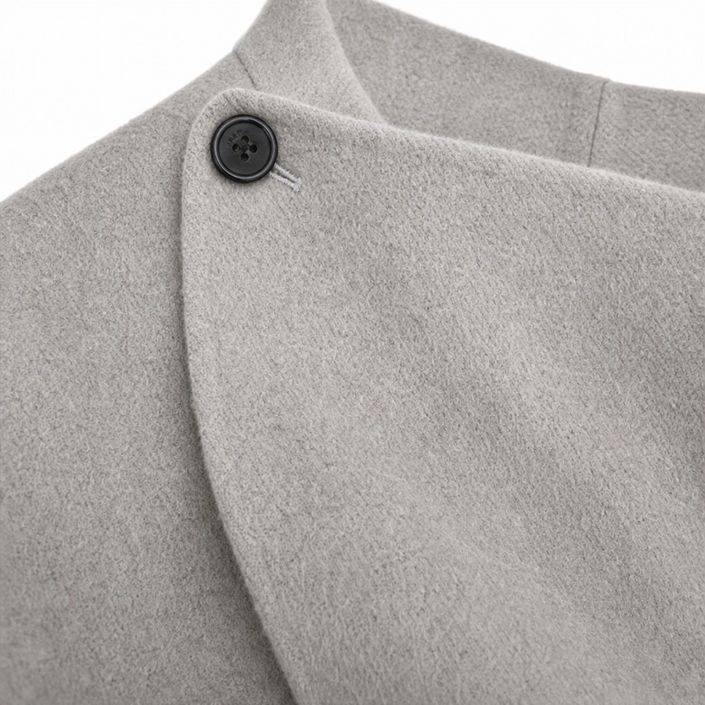 Detalle del botón oscuro y del cierre lateral junto al cuello drapeado de la capa chaqueta gris claro.