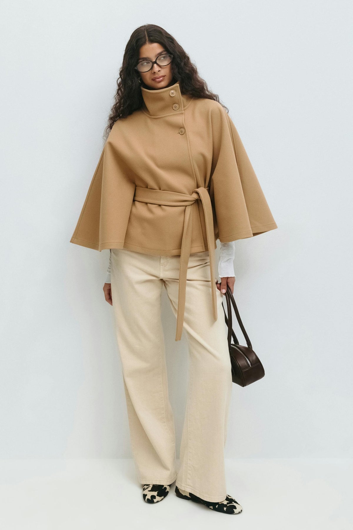 Capa camel llevada con pantalones claros y bolso, mostrando su silueta corta, mangas amplias y cinturón de tiras largas.