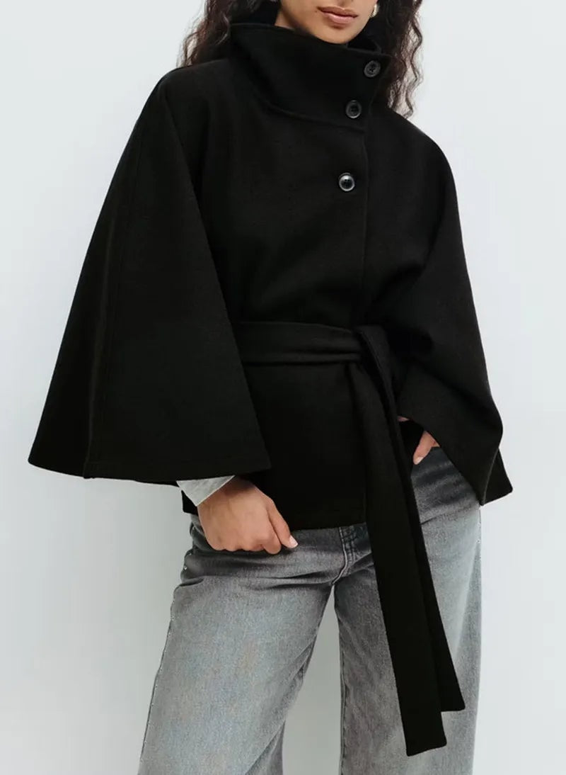 Capa corta negra con cuello alto abotonado, mangas amplias y cinturón largo, combinada con vaqueros grises.