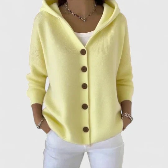 Cárdigan amarillo claro con capucha, mangas largas y fila de botones oscuros, combinado con pantalón blanco.
