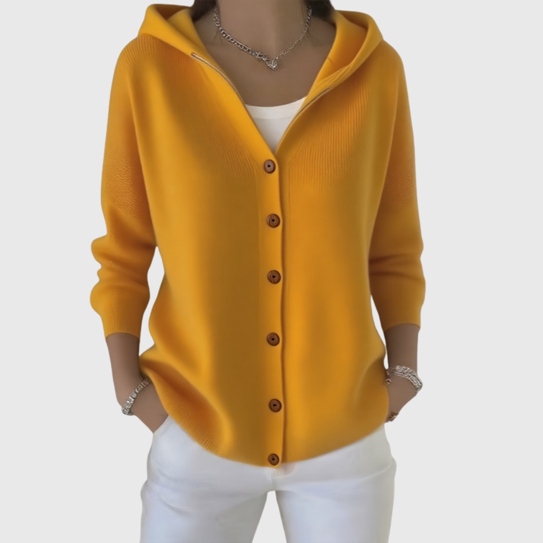 Cárdigan mostaza con capucha y seis botones marrones visibles, combinado con pantalón blanco.