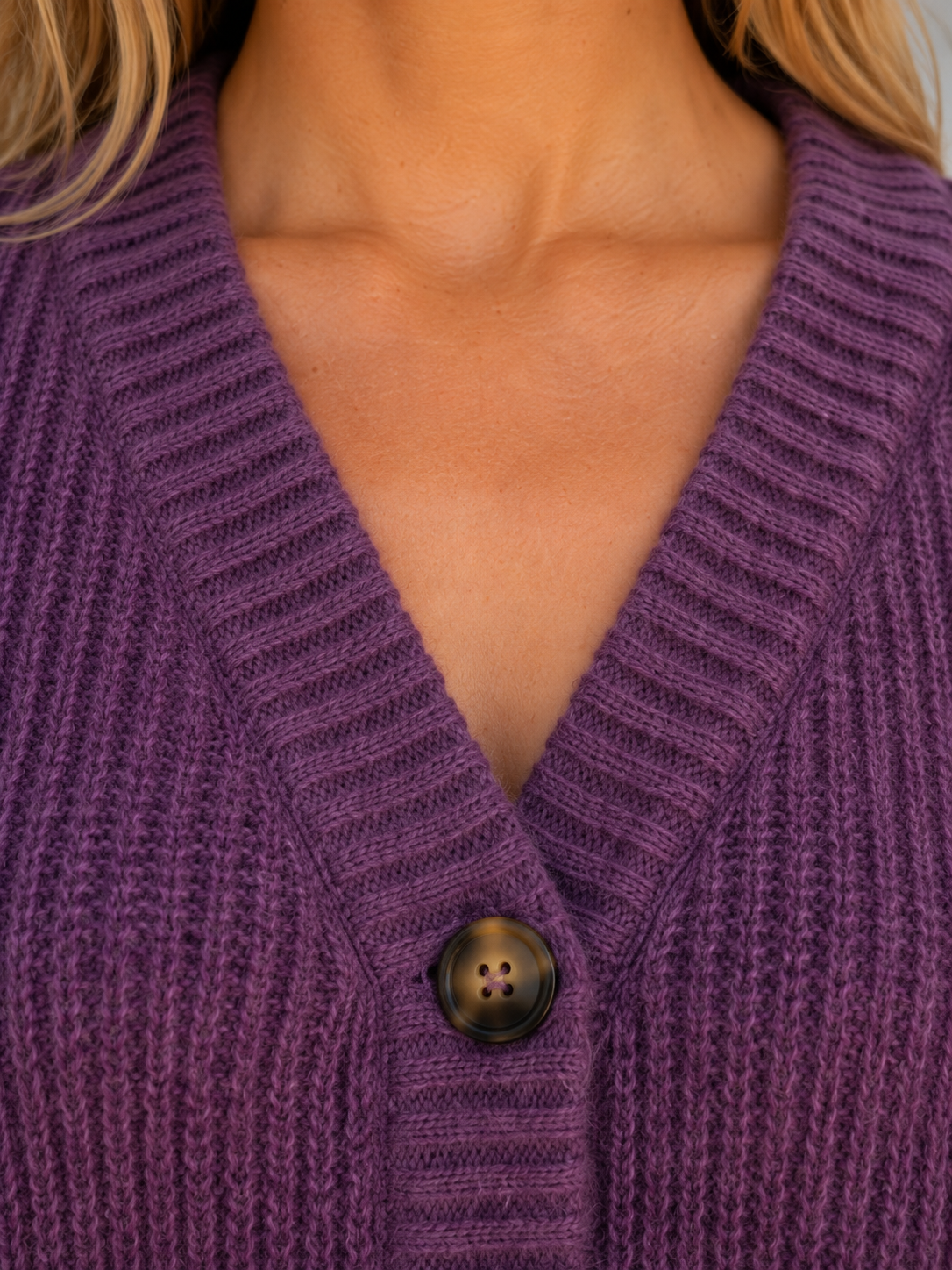 Detalle del cuello de pico y del punto grueso del cárdigan morado, con un botón frontal destacado.