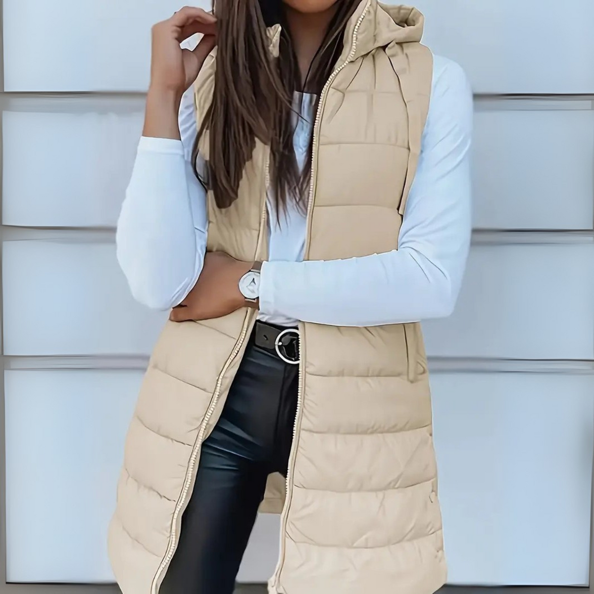 Chaleco acolchado largo beige con capucha y cierre frontal, visto de frente en un conjunto informal.