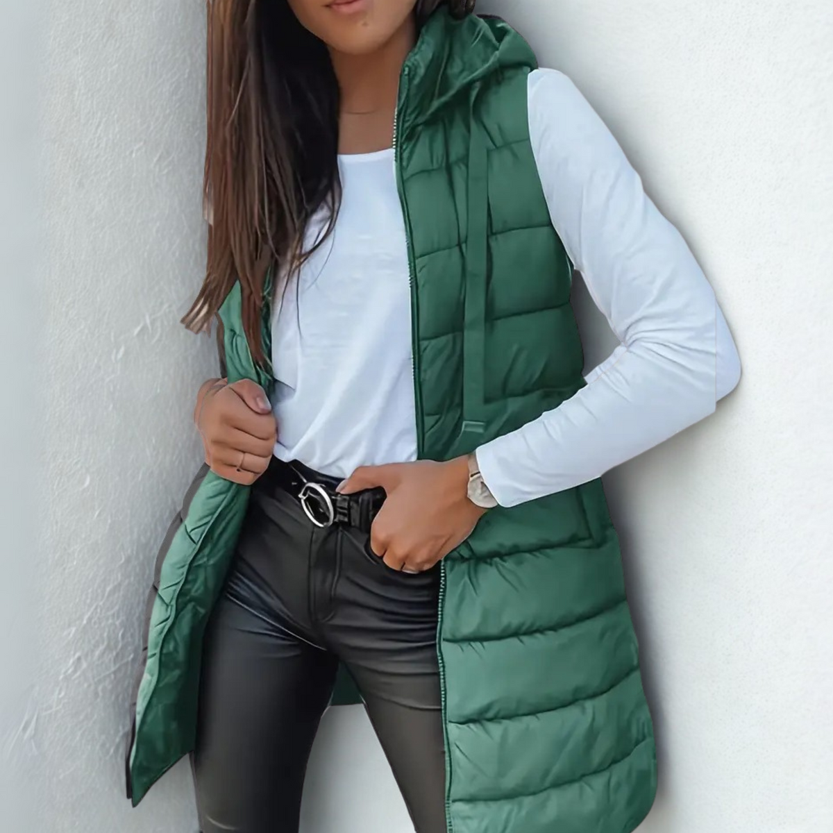 Chaleco acolchado largo verde oscuro con capucha y cierre frontal, combinado con camiseta blanca y pantalones negros.