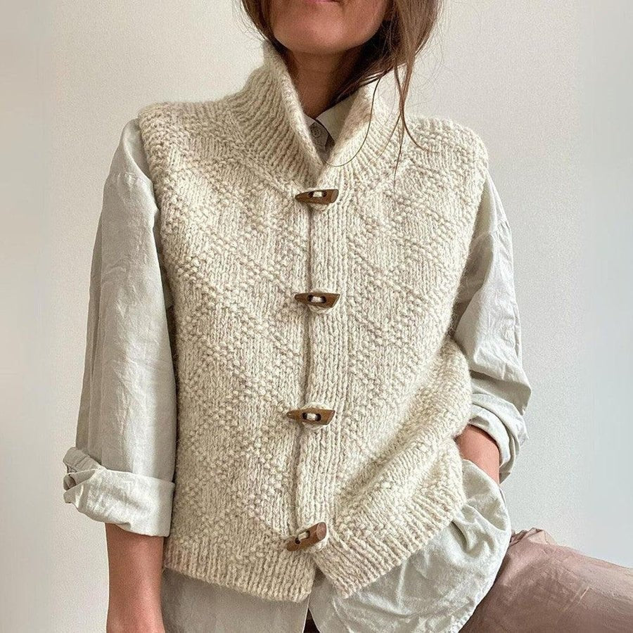 Chaleco beige de punto grueso con cuello alto, dibujo de rombos y cuatro alamares, combinado con camisa blanca.