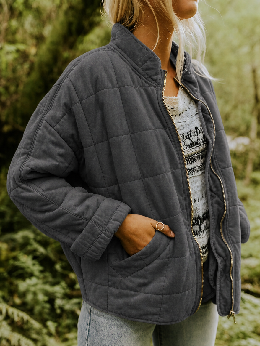 Vista lateral de la chaqueta acolchada gris con cuello alto, cremallera abierta y mano dentro del bolsillo delantero.