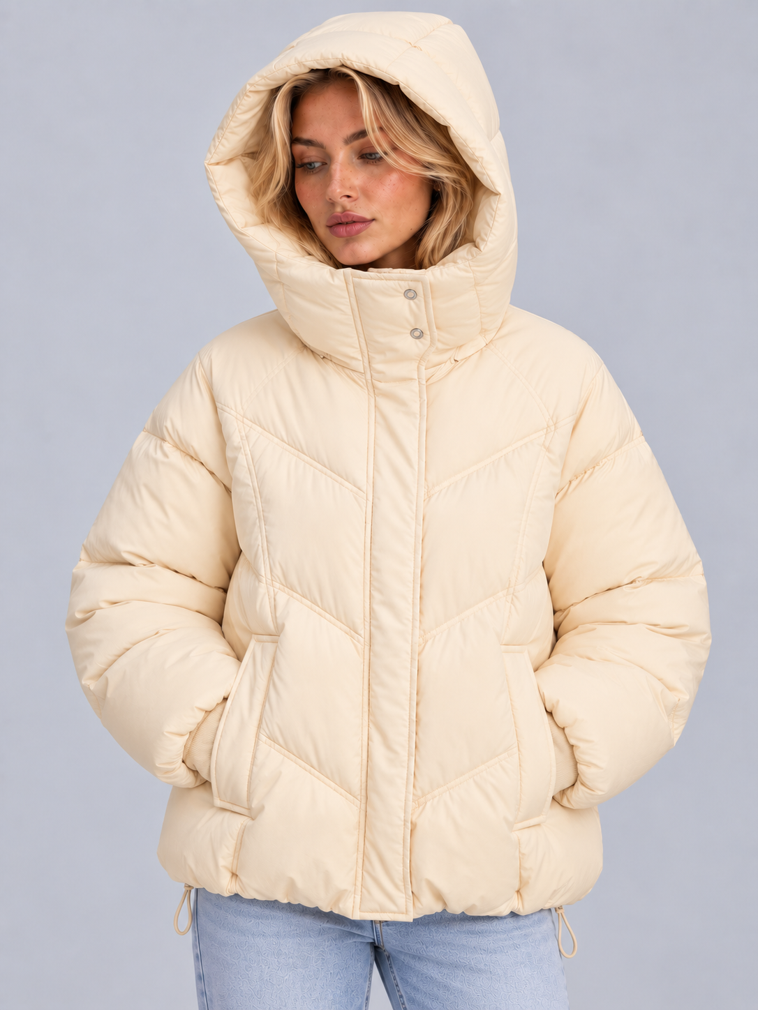 Vista frontal de la chaqueta acolchada crema con la capucha puesta, cuello elevado y ambas manos dentro de los bolsillos delanteros.