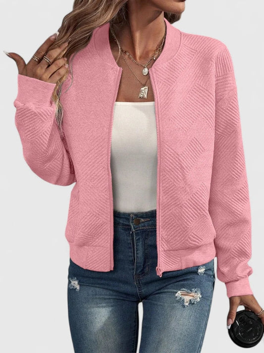 Chaqueta bomber rosa con textura geométrica, cuello redondo, cierre frontal y bolsillos laterales, combinada con vaqueros.