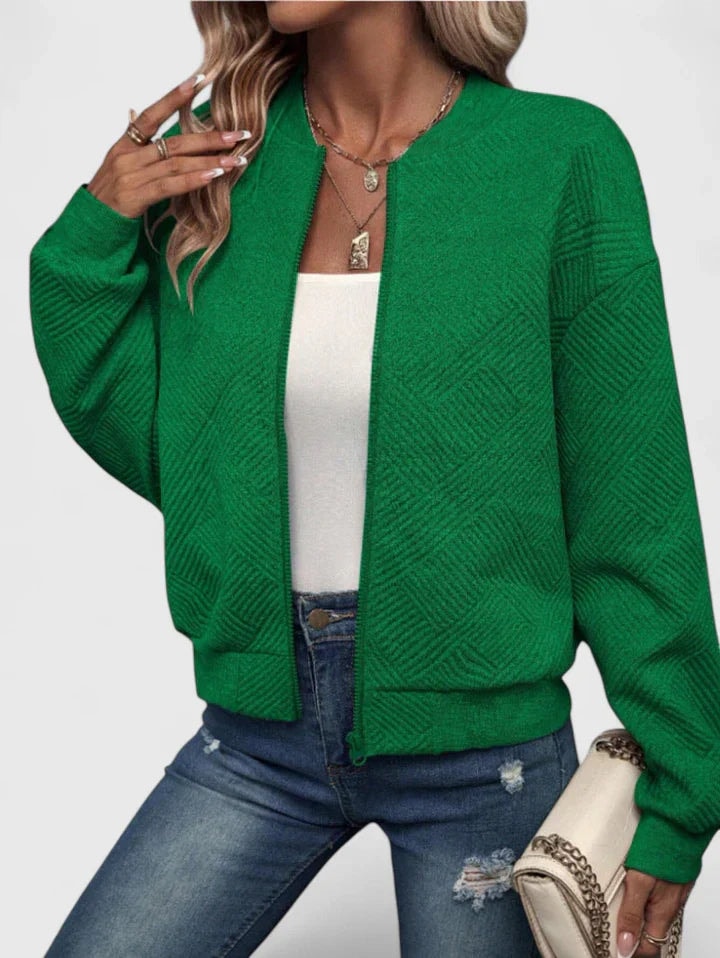 Chaqueta bomber verde con textura geométrica marcada, cierre frontal y bolsillos laterales, combinada con vaqueros y bolso claro.