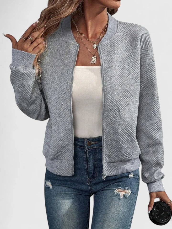 Chaqueta bomber gris claro con patrón geométrico, cuello redondo y cierre frontal, combinada con vaqueros desgastados.