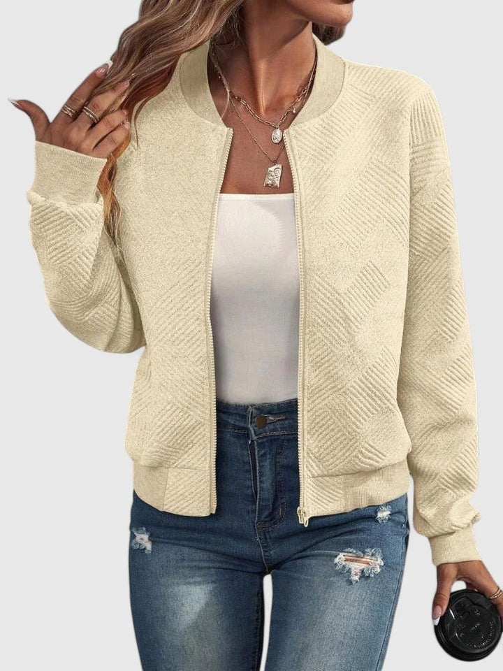 Chaqueta bomber color crema con diseño geométrico, cuello redondo, puños ajustados y cierre frontal, combinada con vaqueros.