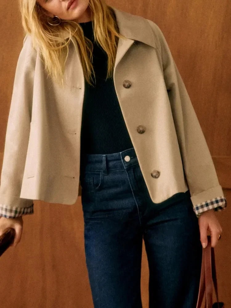 Chaqueta corta beige con cuello camisero, cierre frontal de botones y puños vueltos con detalle de cuadros, combinada con jersey negro y vaqueros oscuros.