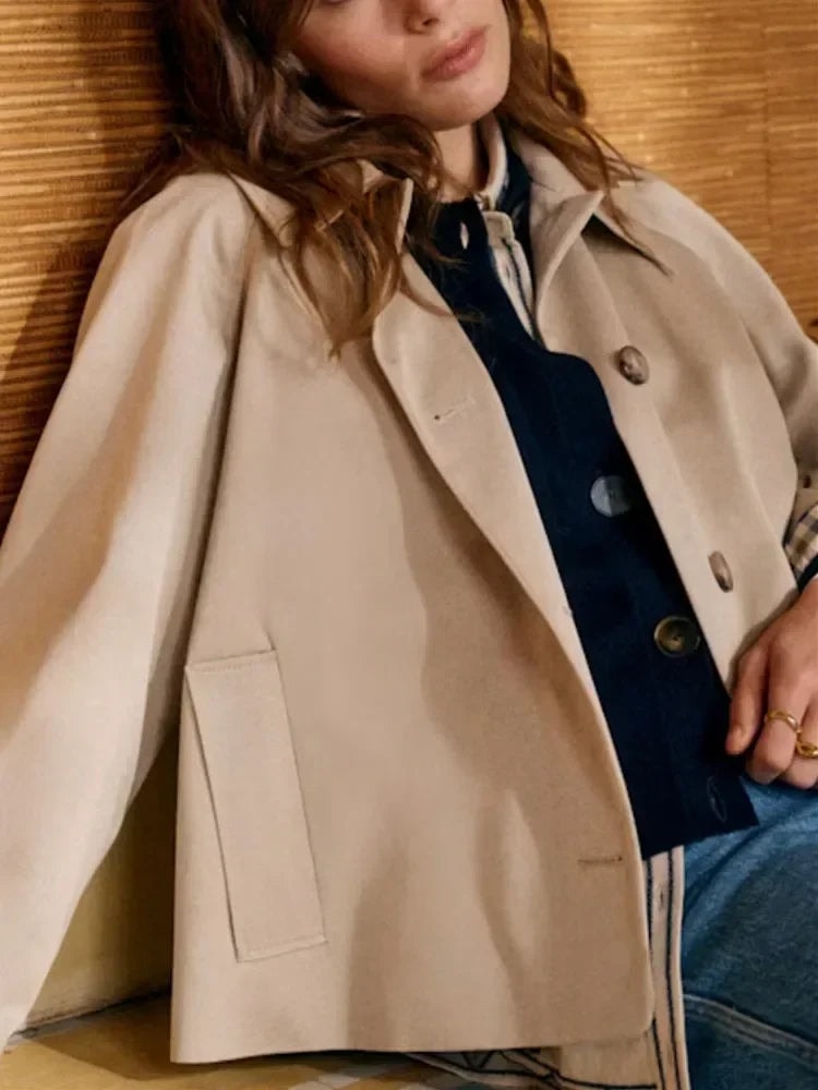 Primer plano lateral de la chaqueta beige abierta, que muestra el bolsillo aplicado, el cuello camisero y el cierre frontal sobre una prenda oscura.