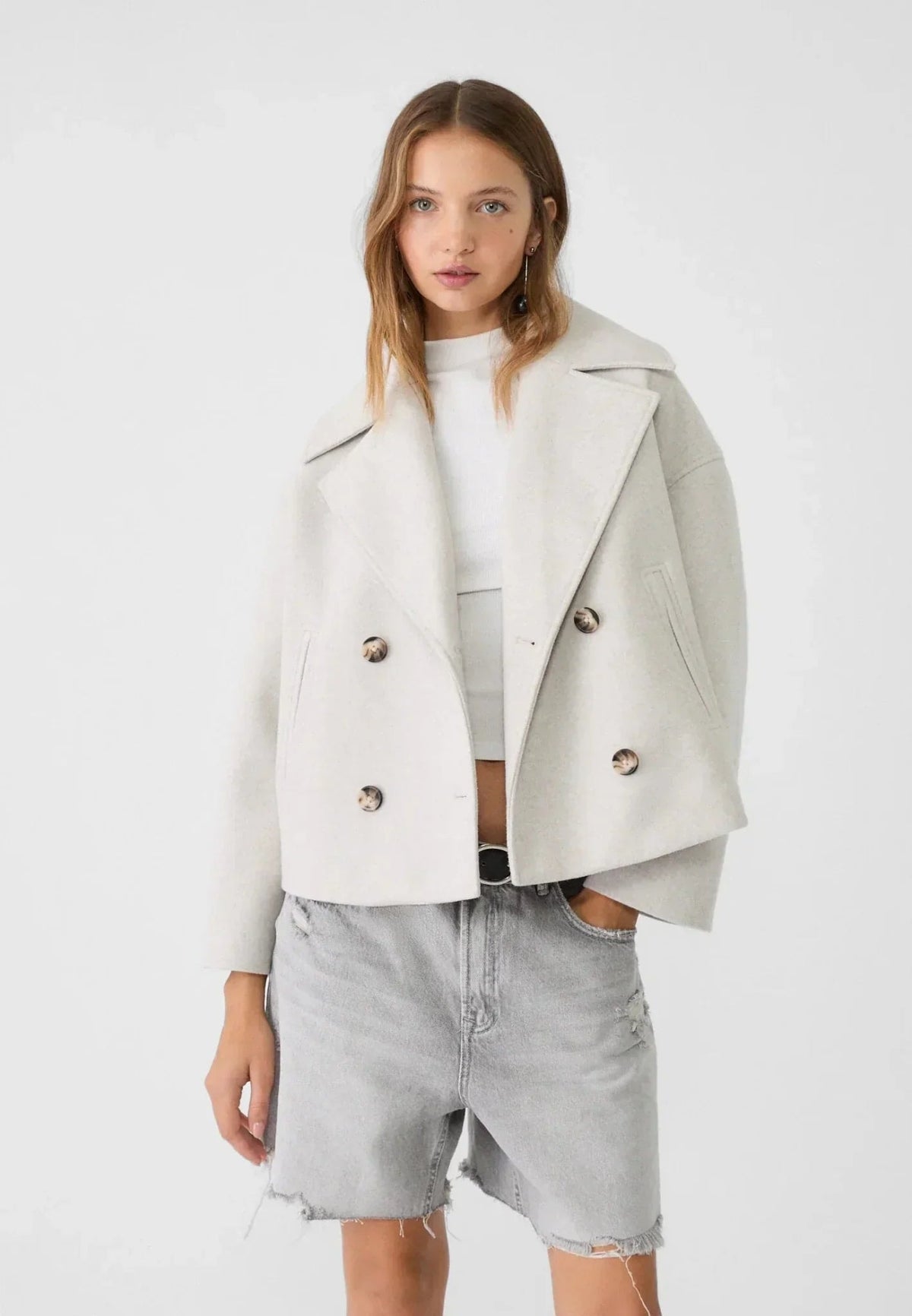 Modelo llevando la chaqueta corta marfil abierta sobre un top claro y pantalones cortos vaqueros.