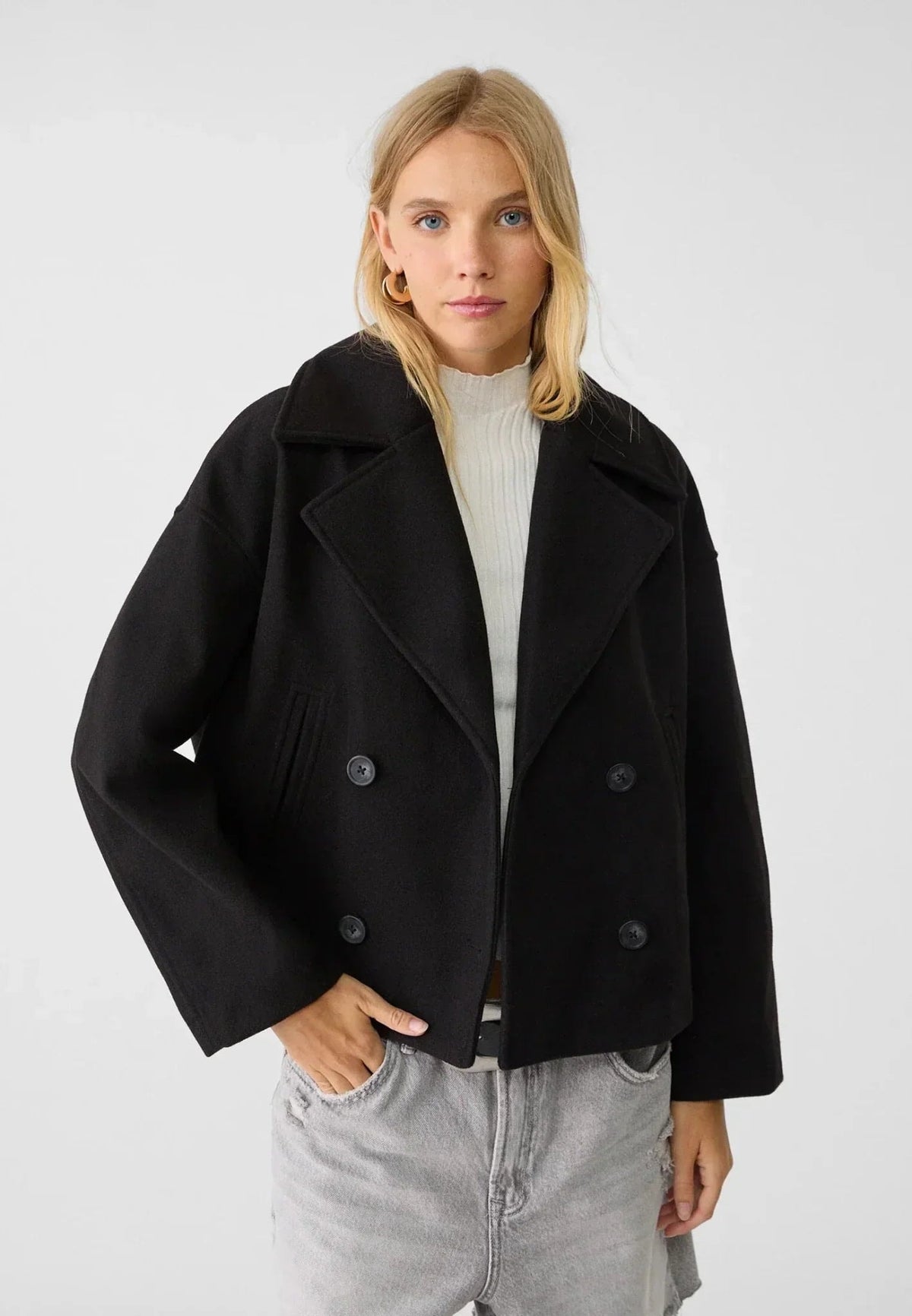Modelo llevando la chaqueta corta negra cerrada, con solapas marcadas y cuatro botones frontales.