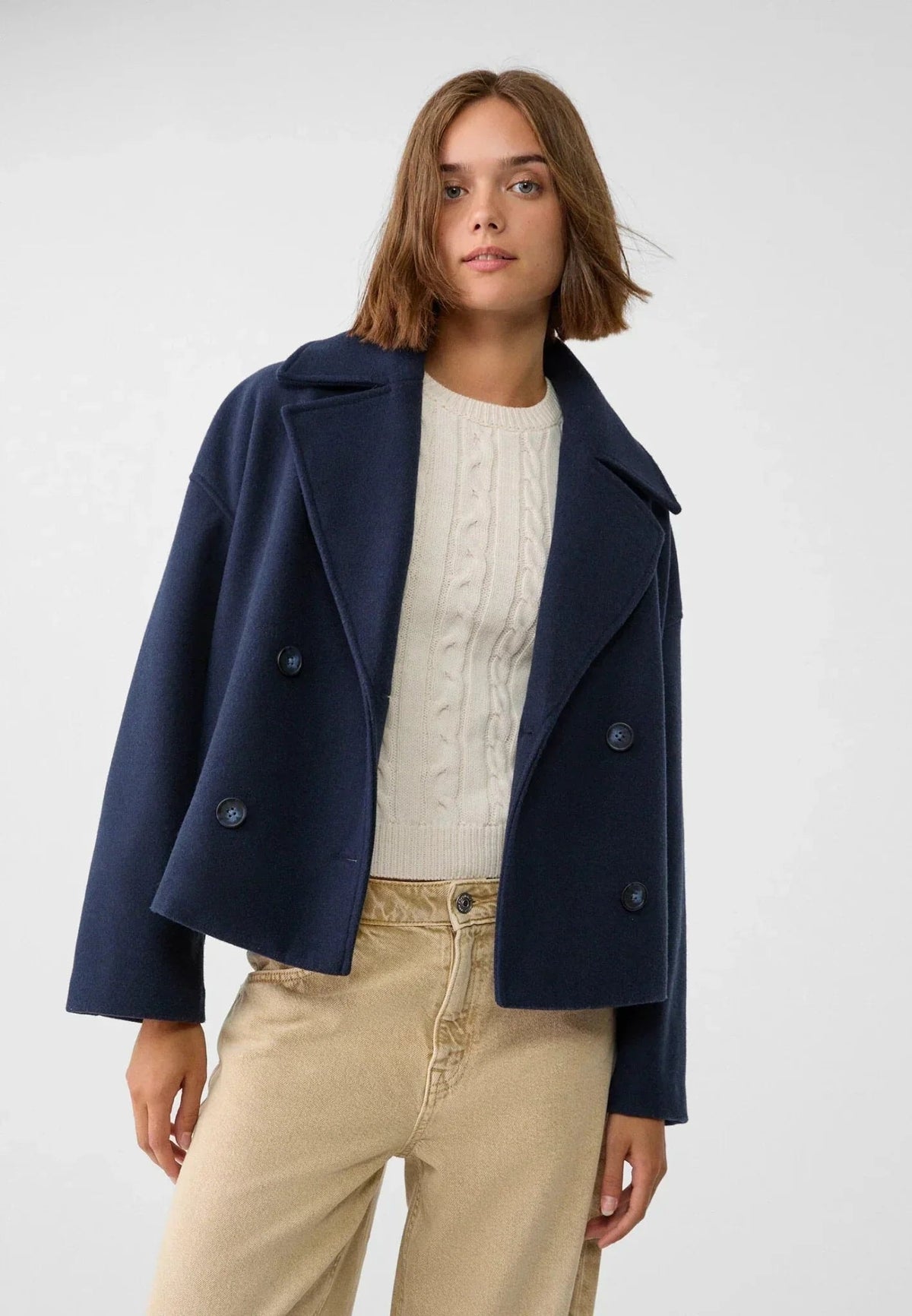 Modelo con chaqueta corta azul marino sobre un jersey claro y pantalones beige.