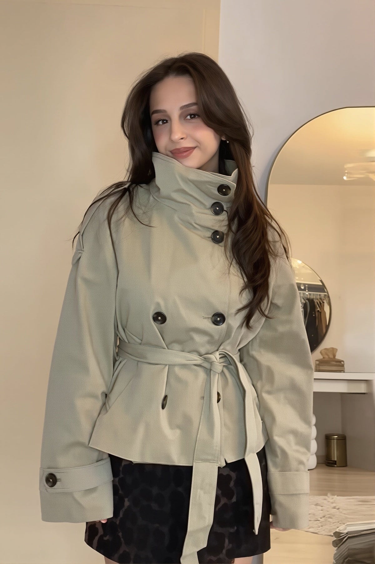 Gabardina corta cruzada en tono claro con cuello alto, botones oscuros, cinturón ceñido y puños abotonados, combinada con pantalón corto estampado.