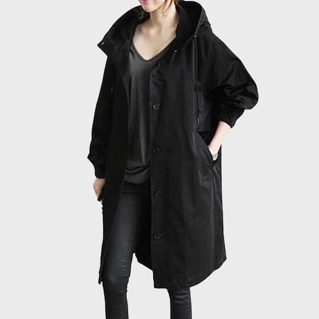 Gabardina larga negra con capucha, botones frontales y bolsillos laterales, combinada con pantalones oscuros.