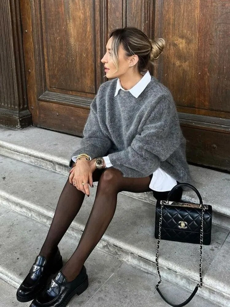 Jersey gris holgado de cuello redondo combinado con una camisa blanca de cuello visible, falda corta y medias oscuras.