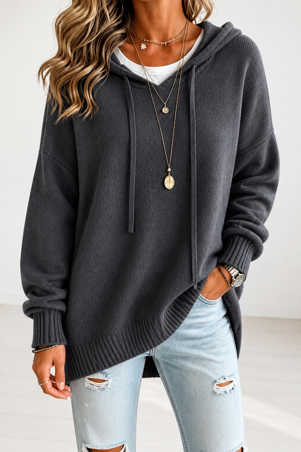 Jersey oversize gris oscuro con capucha y cordones, combinado con vaqueros claros y mostrado con una mano en el bolsillo.