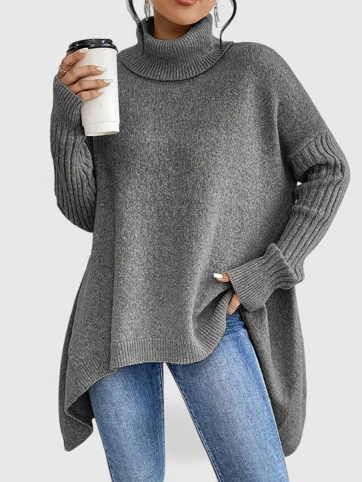 Jersey oversize gris de cuello alto y bajo asimétrico, mostrado de frente con una mano en el bolsillo del vaquero.